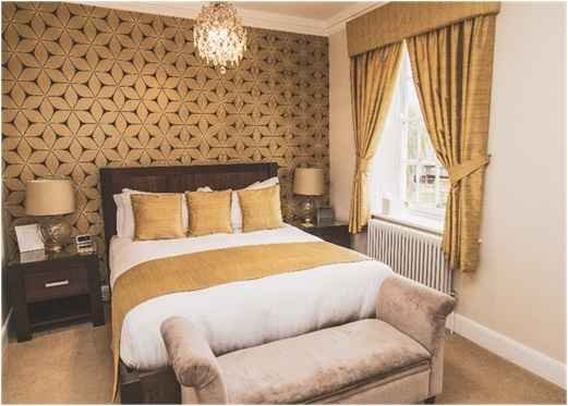 Luxury double bedroom at Downham Hall featuring warm yellow tones, geometric feature wallpaper, king-size bed, and elegant boutique décor.