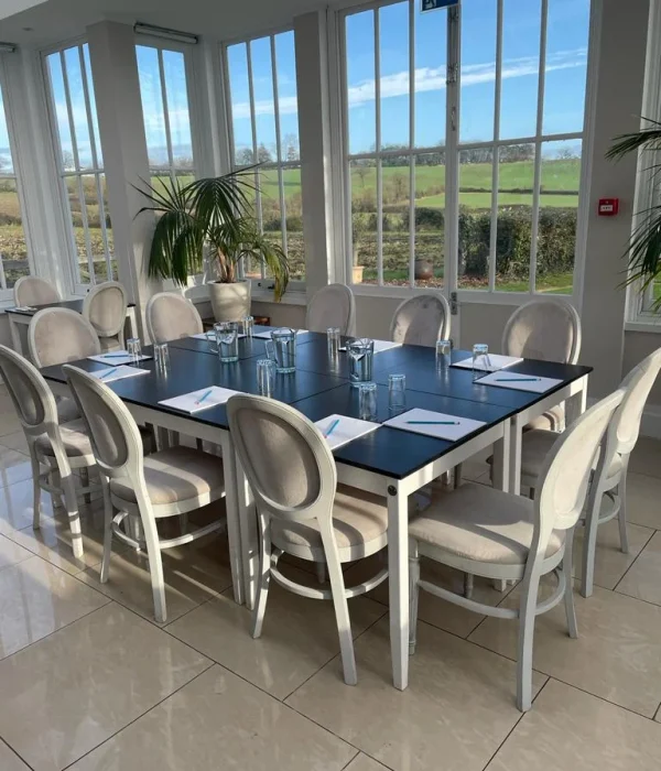 A circular meeting table with white chairs and notebooks under a chandelier in the light-filled Downham Hall Orangery.