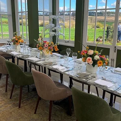 A long marble dining table with floral centerpieces and views of the Essex countryside through large windows in the Orangery.